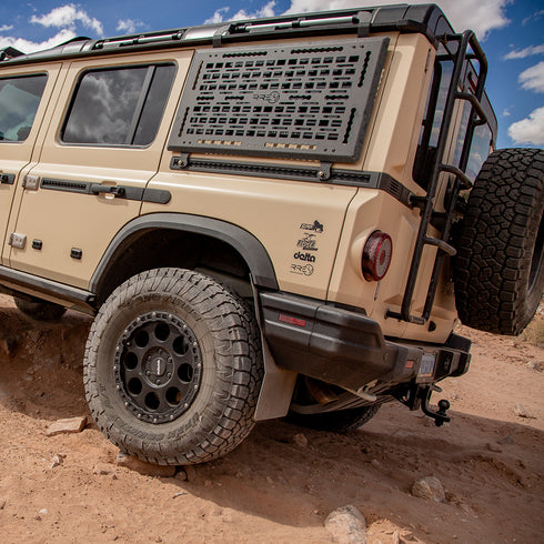 A Black Exterior cargo carrier made by Rough Road Equipment with the RRE logo stamped into it, shown on an Ineos Grenadier. Featuring three different attachment points for various systems like L-track (Airline Track), MOLLE, and Carrage bolt. Show with the included attachment points for the factory grab handles. The included upper MOLLE panel is not shown.
Desert Tan off-road Grenadier on a dirt terrain with a clear sky. Delta Wheels, Reiger V5 Suspension