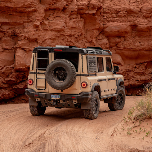 A Black Exterior cargo carrier made by Rough Road Equipment with the RRE logo stamped into it, shown on an Ineos Grenadier. Featuring three different attachment points for various systems like L-track (Airline Track), MOLLE, and Carrage bolt. Show with the included attachment points for the factory grab handles. The included upper MOLLE panel is not shown.
Beige off-road vehicle with a spare tire on a dirt road against a rocky cliff.
Vehicle shown is equipped with Delta Wheels & the Reiger V5 Suspension