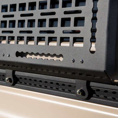 Close-up of a black metal grille or cover on a vehicle, with a beige background. A Black Exterior cargo carrier made by Rough Road Equipment with the RRE logo stamped into it, shown on an Ineos Grenadier. Featuring three different attachment points for various systems like L-track (Airline Track), MOLLE, and Carrage bolt. Showing the attachment to the factory L-Track on the Grenadiers side panels.