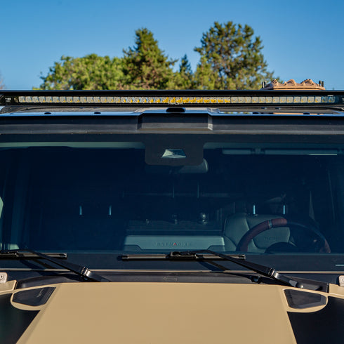 Front view of an Ineos Grenadier with an LED Lite bar and windshield wipers, set against a clear blue sky in a forest.