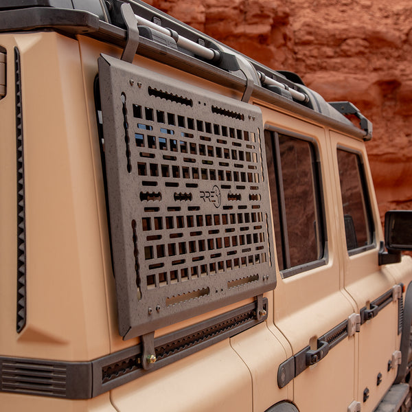 Beige off-road vehicle with a black grille guard in a desert setting. A Black Exterior cargo carrier made by Rough Road Equipment with the RRE logo stamped into it, shown on an Ineos Grenadier. Featuring three different attachment points for various systems like L-track (Airline Track), MOLLE, and Carrage bolt. Show with the included attachment points for the factory grab handles. The included upper MOLLE panel is not shown. 