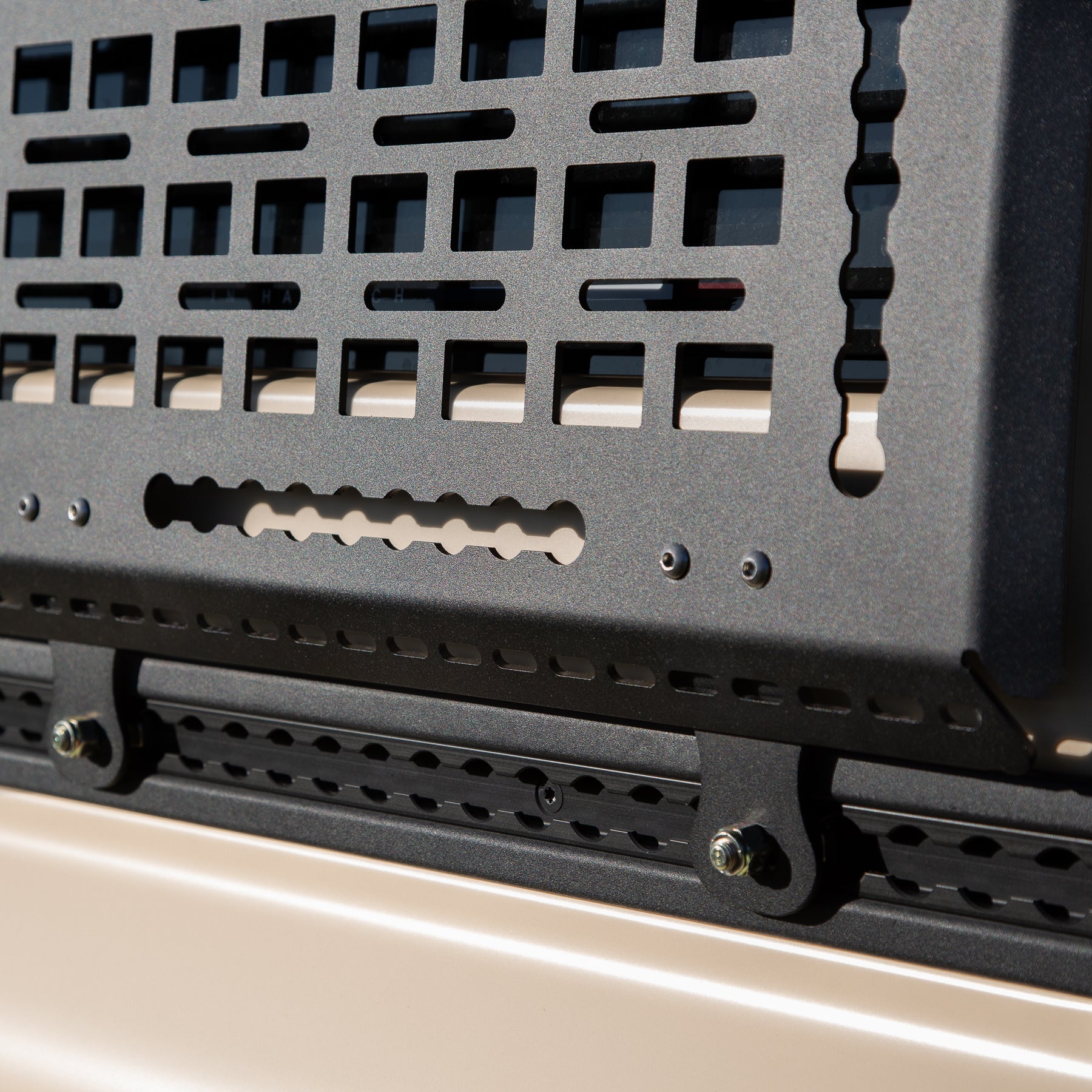 Close-up of a black metal grille or cover on a vehicle, with a beige background. A Black Exterior cargo carrier made by Rough Road Equipment with the RRE logo stamped into it, shown on an Ineos Grenadier. Featuring three different attachment points for various systems like L-track (Airline Track), MOLLE, and Carrage bolt. Showing the attachment to the factory L-Track on the Grenadiers side panels. 