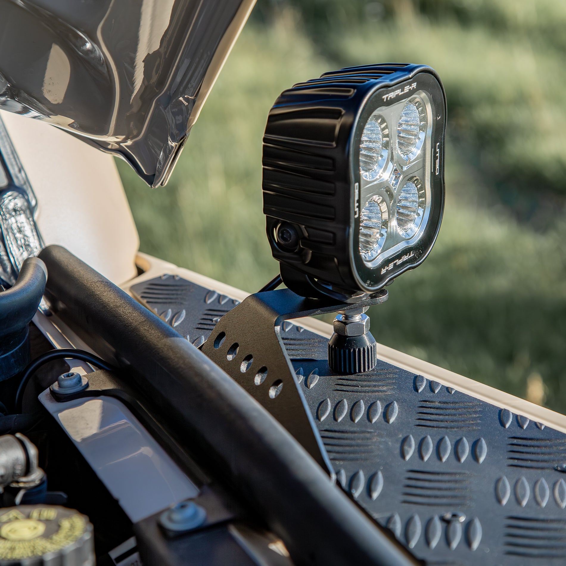 A powder-coated black aluminum Ditch Light Bracket and LED Triple-R Brand Offroad LED light mounted on the edge of the hood with no drilling needed, showing a Spacer to reduce vibration and bouncing of the light mounted on the Ineos Grenadier or Quartermaster.  This image Shows how the Bracket attaches to Factory screw mounts and goes under the existing Bulb seal
