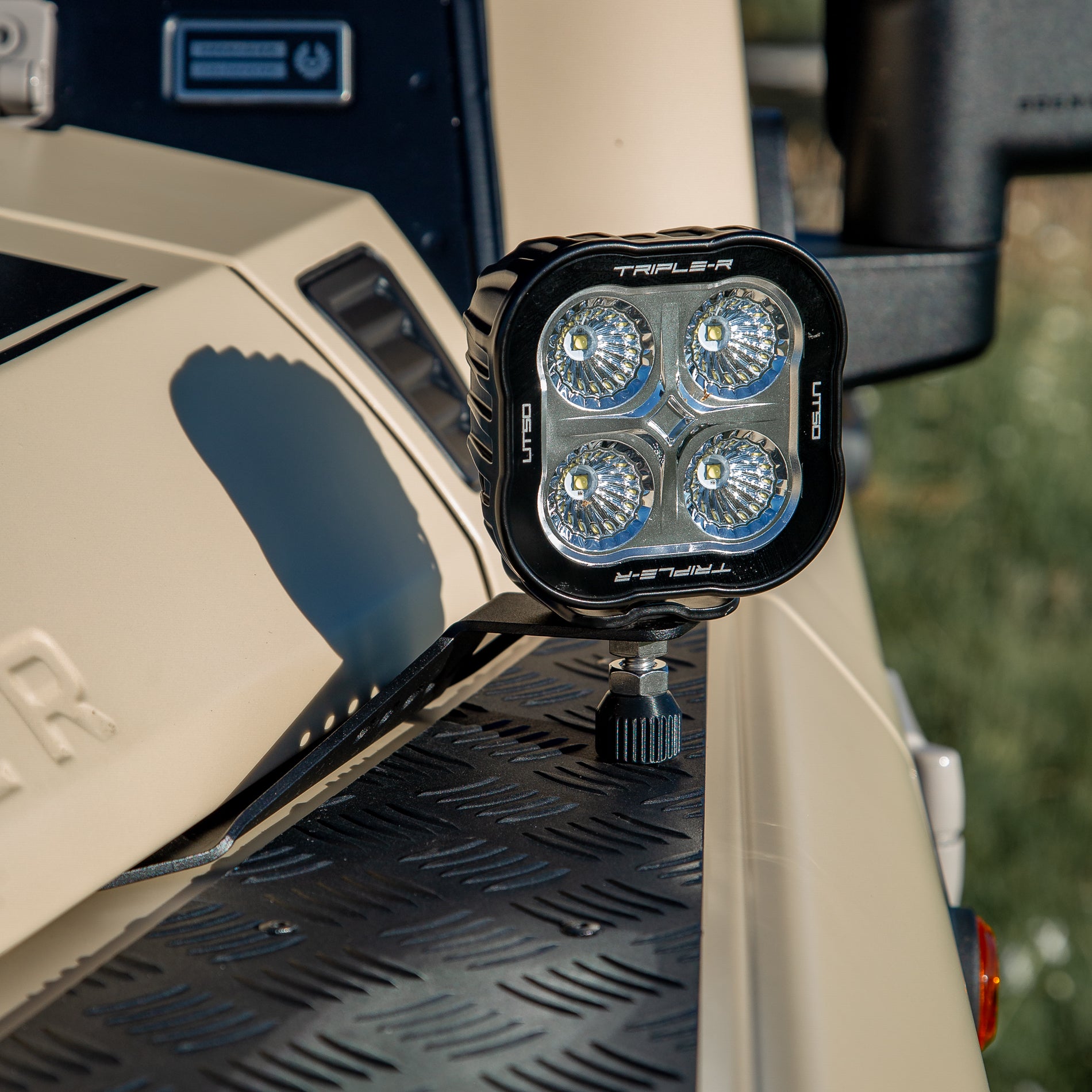 A powder-coated black aluminum Ditch Light Bracket and LED Triple-R Brand Offroad LED light mounted on the edge of the hood with no drilling needed, showing a Spacer to reduce vibration and bouncing of the light mounted on the Ineos Grenadier or Quartermaster. 

Triple R Offroad light mounted on a vehicle with a blurred natural background