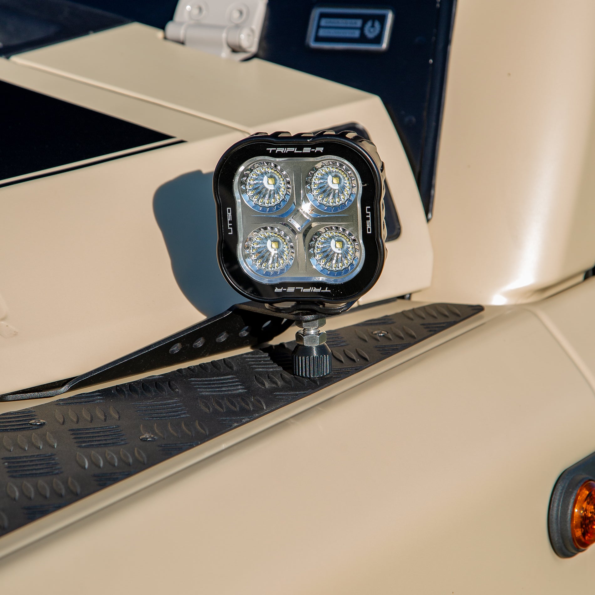 A powder-coated black aluminum Ditch Light Bracket and LED Triple-R Brand Offroad LED light mounted on the edge of the hood with no drilling needed, showing a Spacer to reduce vibration and bouncing of the light mounted on the Ineos Grenadier or Quartermaster. 
Off-road vehicle with a mounted light bar on a Desert Tan background