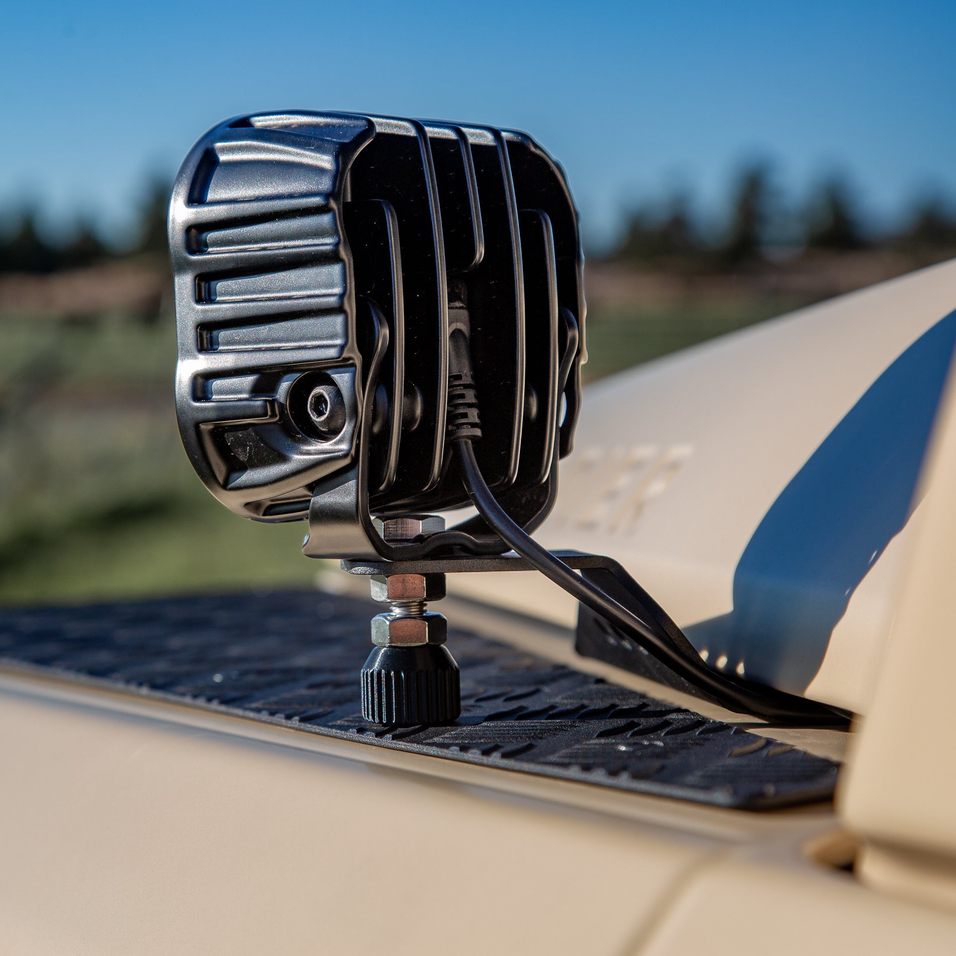A powder-coated black aluminum Ditch Light Bracket and LED Triple-R Brand Offroad LED light mounted on the edge of the hood with no drilling needed, showing a Spacer to reduce vibration and bouncing of the light mounted on the Ineos Grenadier or Quartermaster. 
Black off-road light mounted on a vehicle with a blurred natural background