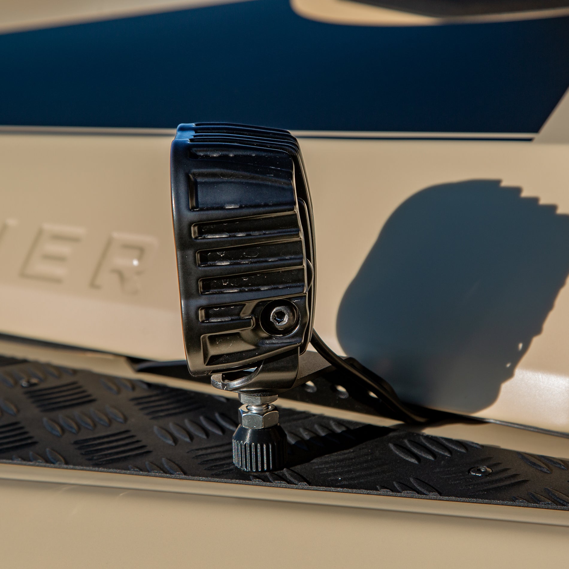 A powder-coated black aluminum Ditch Light Bracket and LED Triple-R Brand Offroad LED light mounted on the edge of the hood with no drilling needed, showing a Spacer to reduce vibration and bouncing of the light mounted on the Ineos Grenadier or Quartermaster. 
Black off-road light mounted on a vehicle with 'GRENADIER' branding.