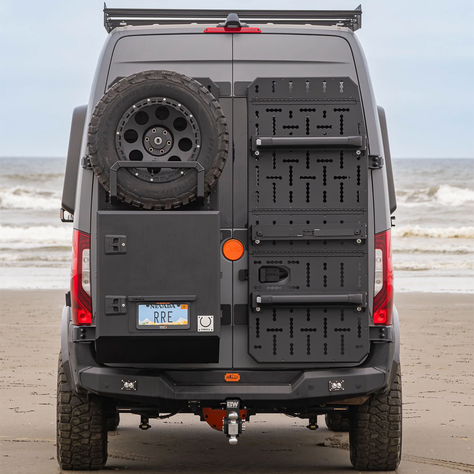 X-Bull aluminum cargo carrier mounted to a Sprinter rear door, holding a full-size spare tire and gear box.