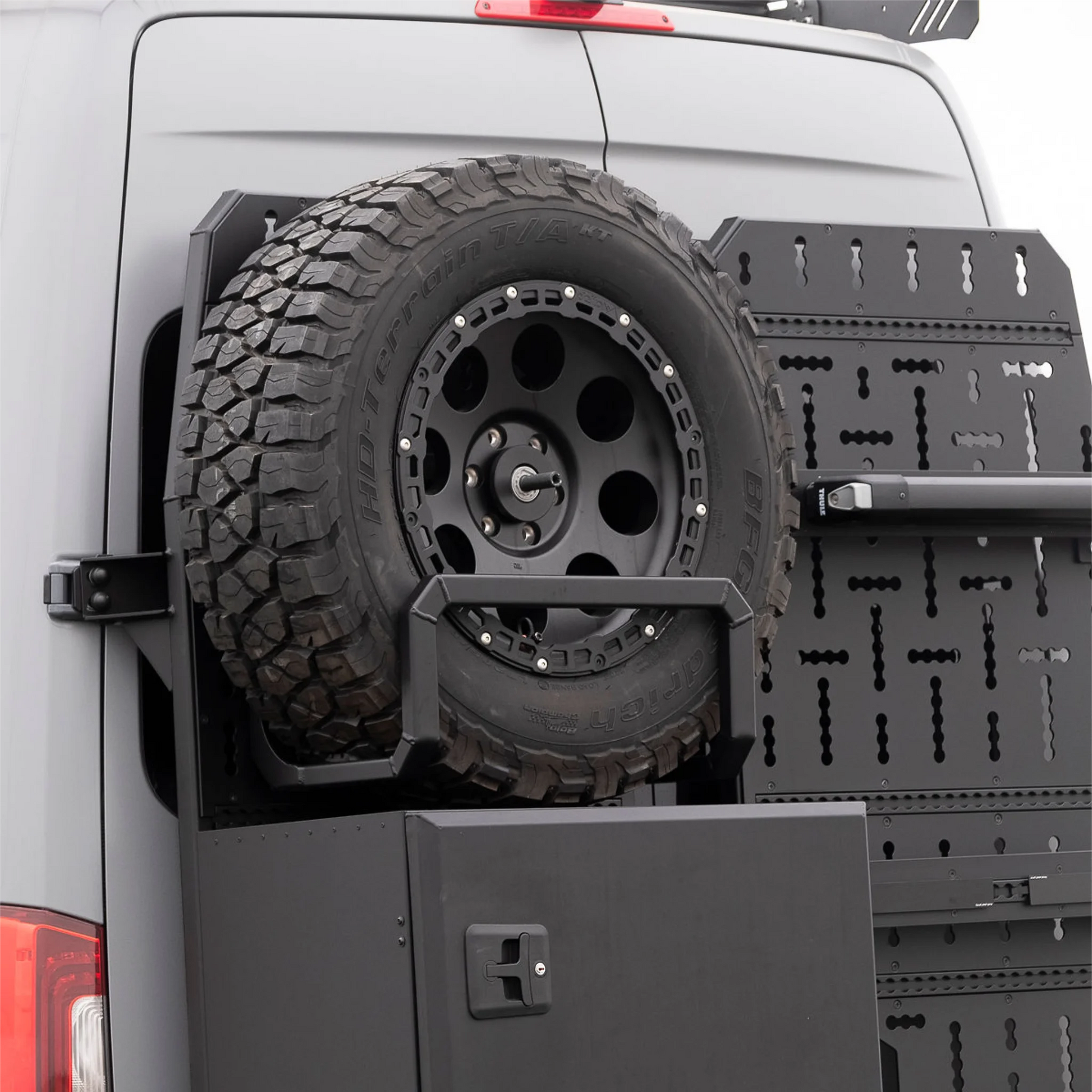 Black aluminum XBULL spare wheel carrier mounted to a rear door cargo rack on a Mercedes Sprinter, holding a full-size off-road tire.