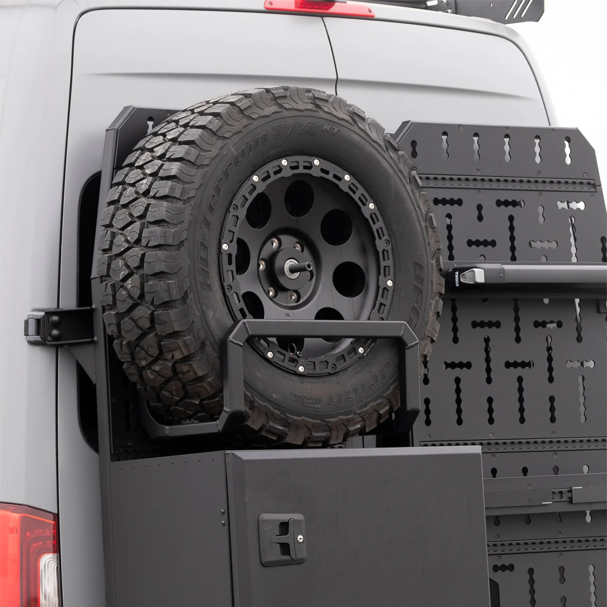 Black aluminum XBULL spare wheel carrier mounted to a rear door cargo rack on a Mercedes Sprinter, holding a full-size off-road tire.