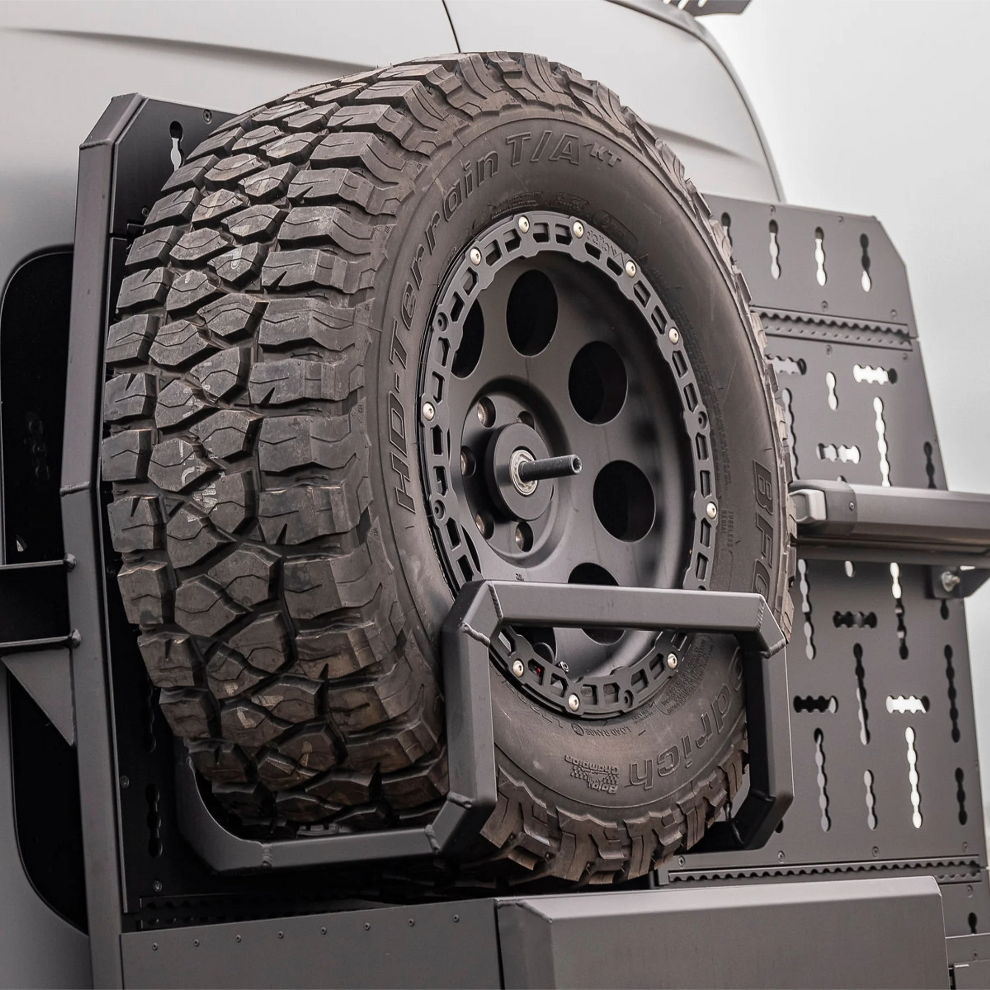 Black aluminum XBULL spare wheel carrier mounted to a rear door cargo rack on a Mercedes Sprinter, holding a full-size off-road tire.