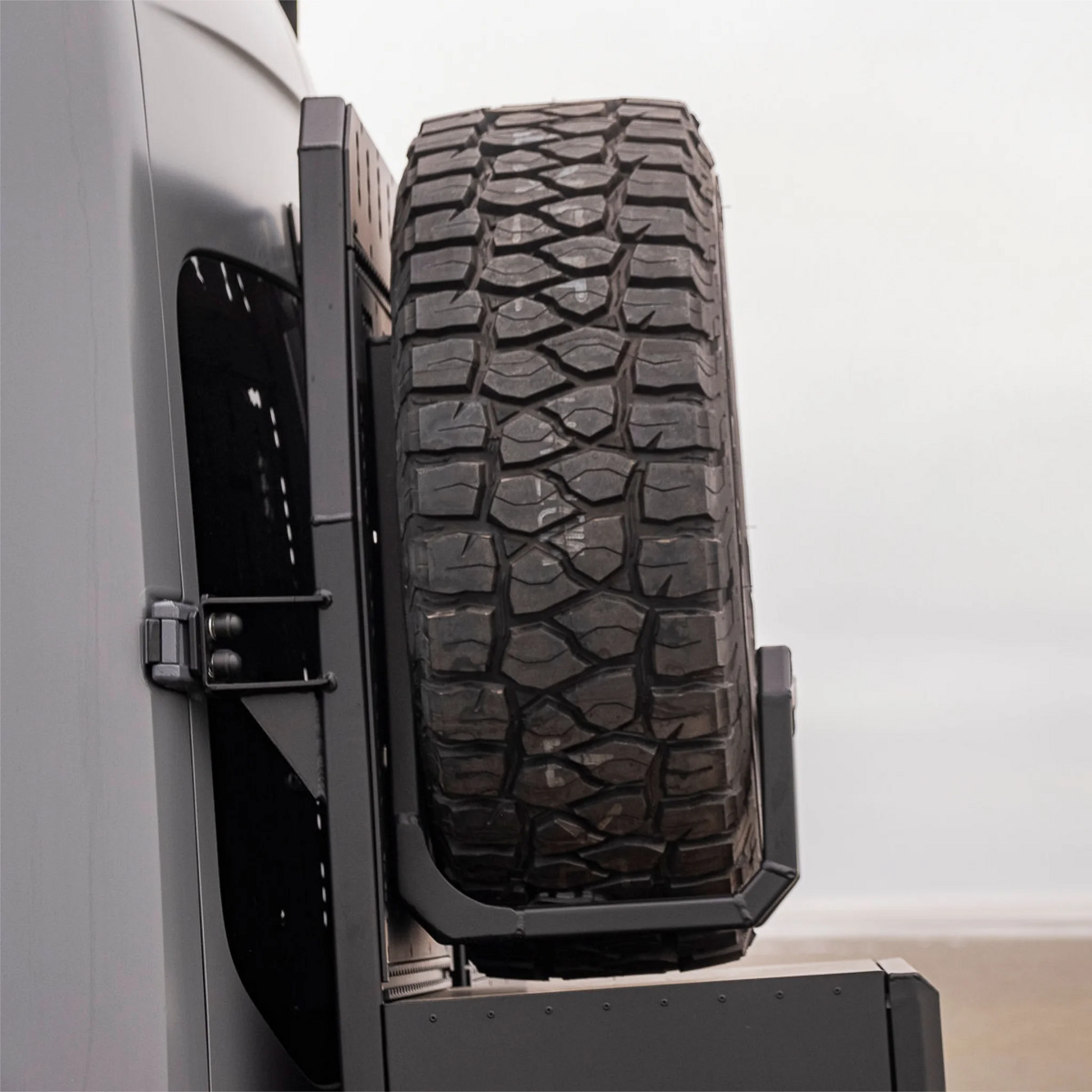 Black aluminum XBULL spare wheel carrier mounted to a rear door cargo rack on a Mercedes Sprinter, holding a full-size off-road tire.