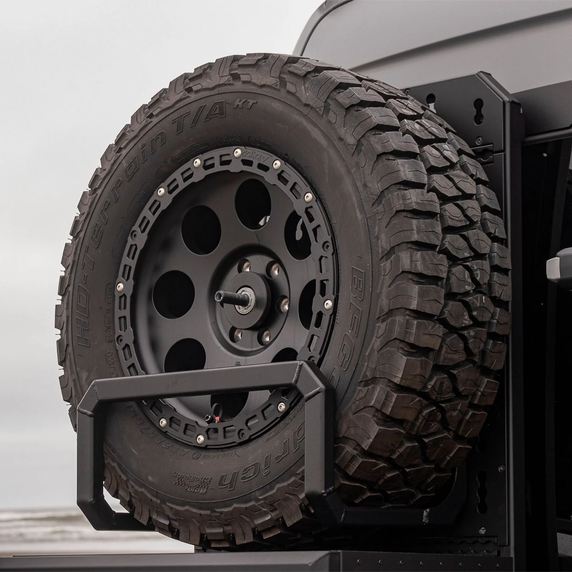 Black aluminum XBULL spare wheel carrier mounted to a rear door cargo rack on a Mercedes Sprinter, holding a full-size off-road tire.