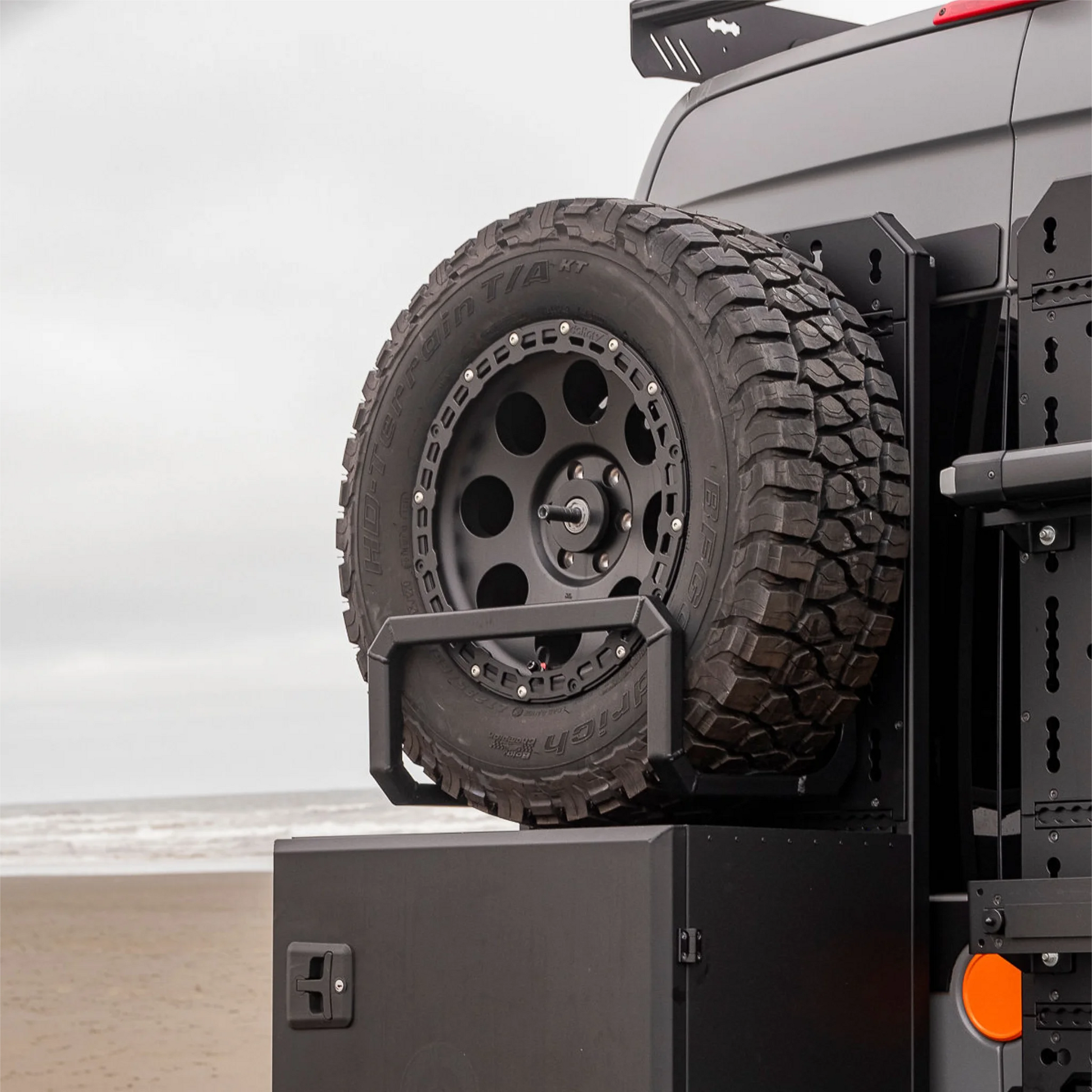 Black aluminum XBULL spare wheel carrier mounted to a rear door cargo rack on a Mercedes Sprinter, holding a full-size off-road tire.