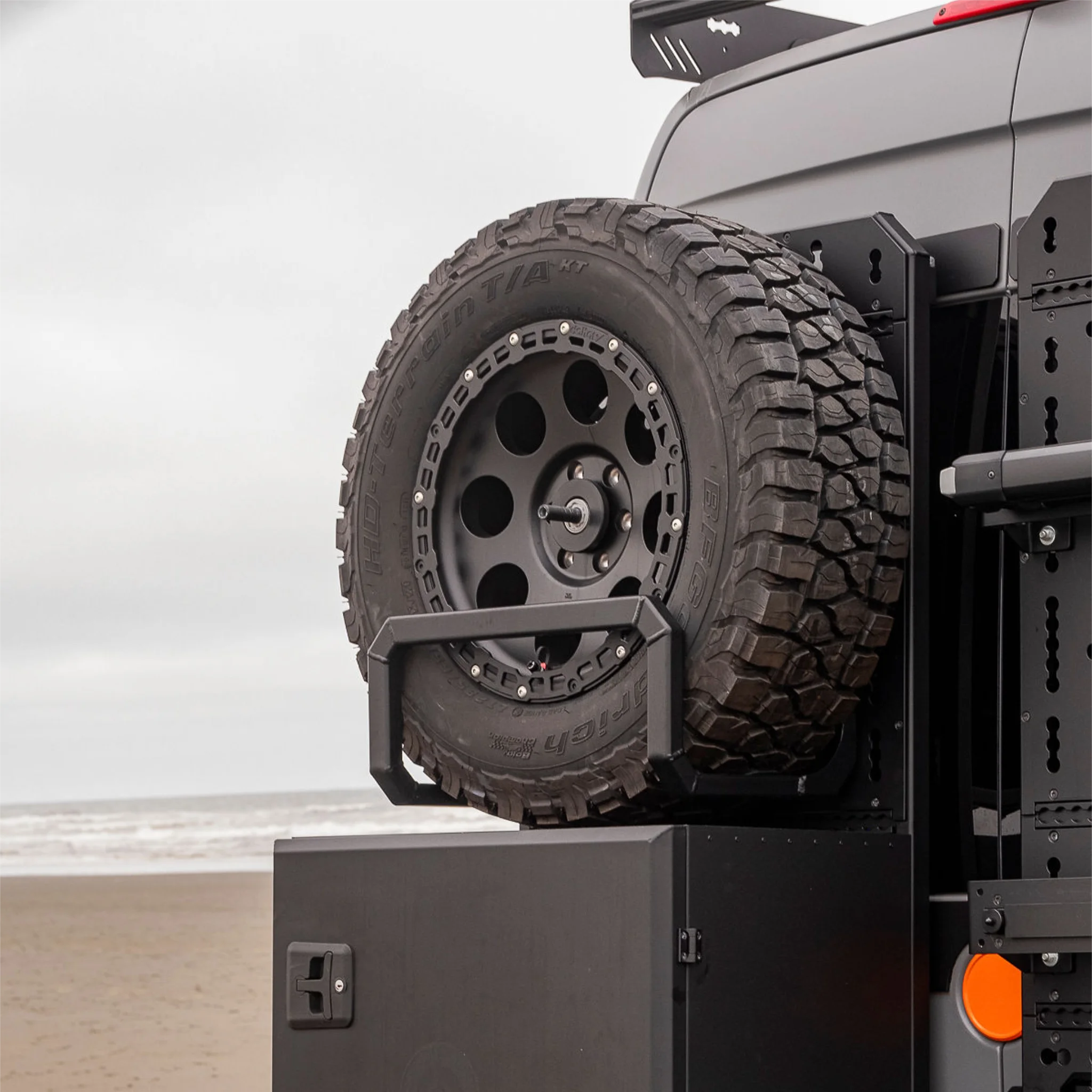 Black aluminum XBULL spare wheel carrier mounted to a rear door cargo rack on a Mercedes Sprinter, holding a full-size off-road tire.