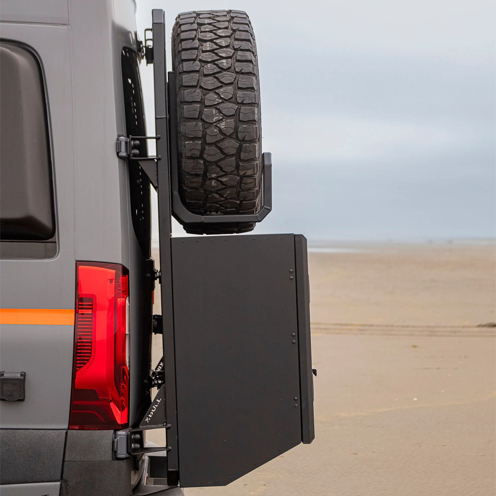 Black aluminum XBULL spare wheel carrier mounted to a rear door cargo rack on a Mercedes Sprinter, holding a full-size off-road tire.
