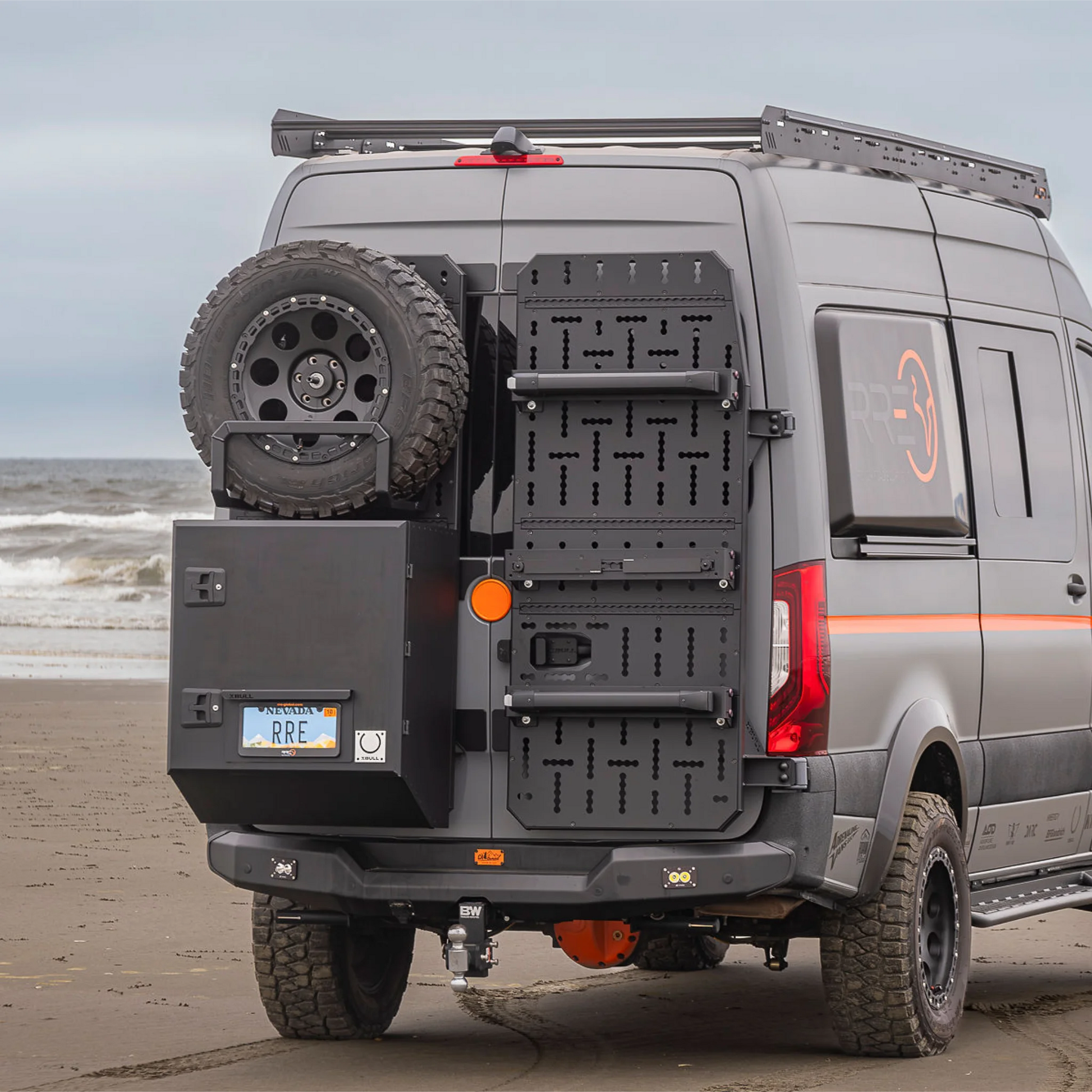 X-Bull aluminum cargo carrier mounted to a Sprinter rear door, holding a full-size spare tire and gear box.