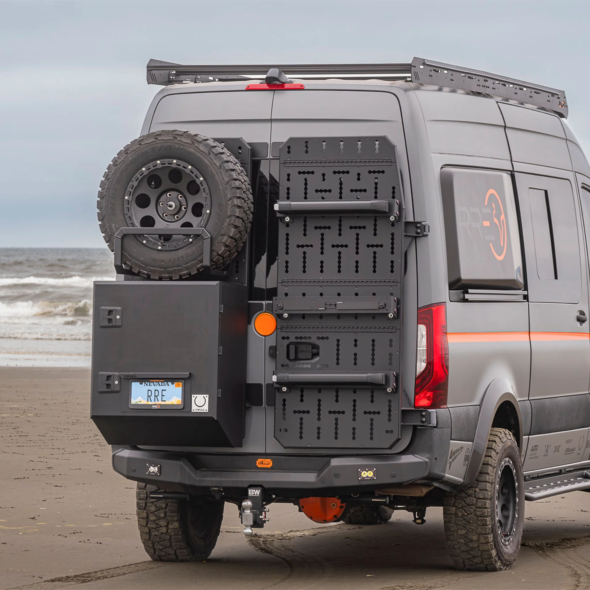 X-Bull aluminum cargo carrier mounted to a Sprinter rear door, holding a full-size spare tire and gear box.
