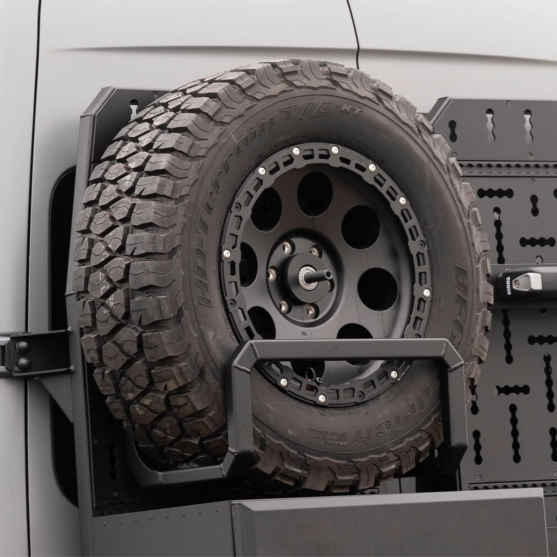 Black aluminum XBULL spare wheel carrier mounted to a rear door cargo rack on a Mercedes Sprinter, holding a full-size off-road tire.