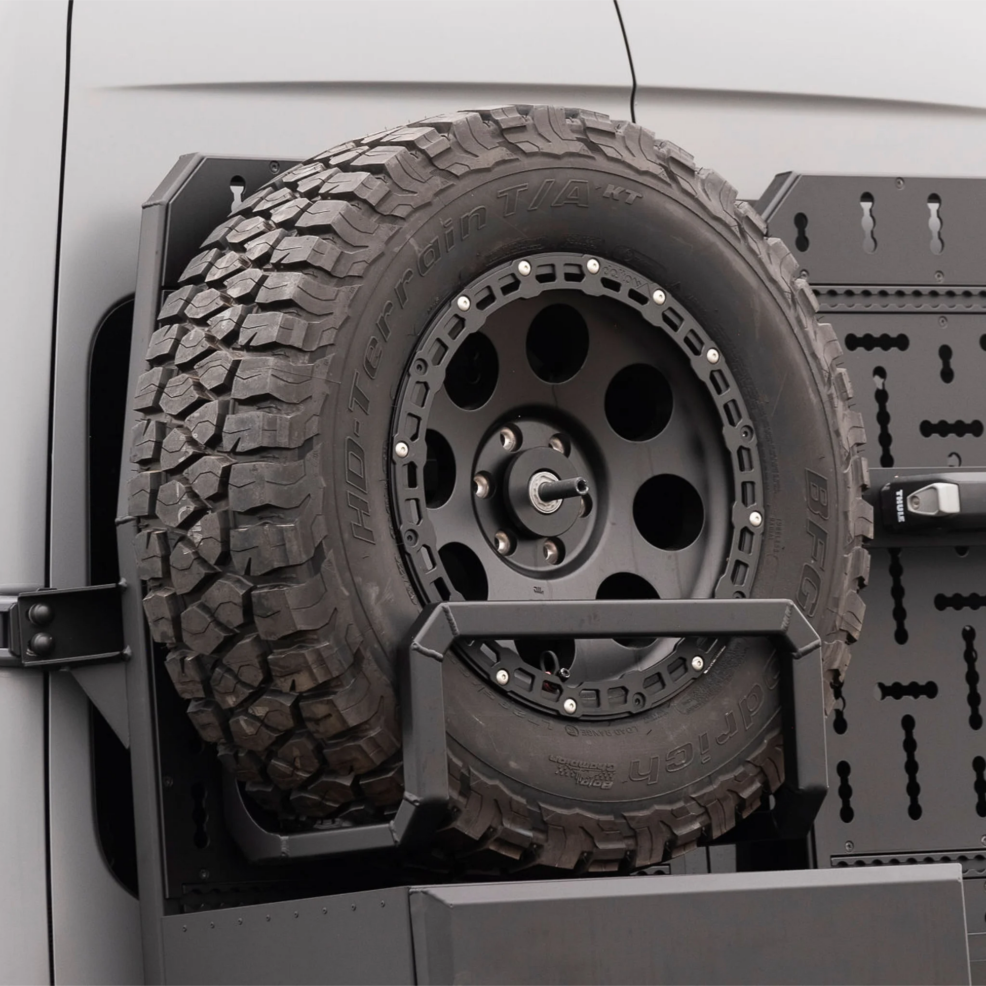 Black aluminum XBULL spare wheel carrier mounted to a rear door cargo rack on a Mercedes Sprinter, holding a full-size off-road tire.